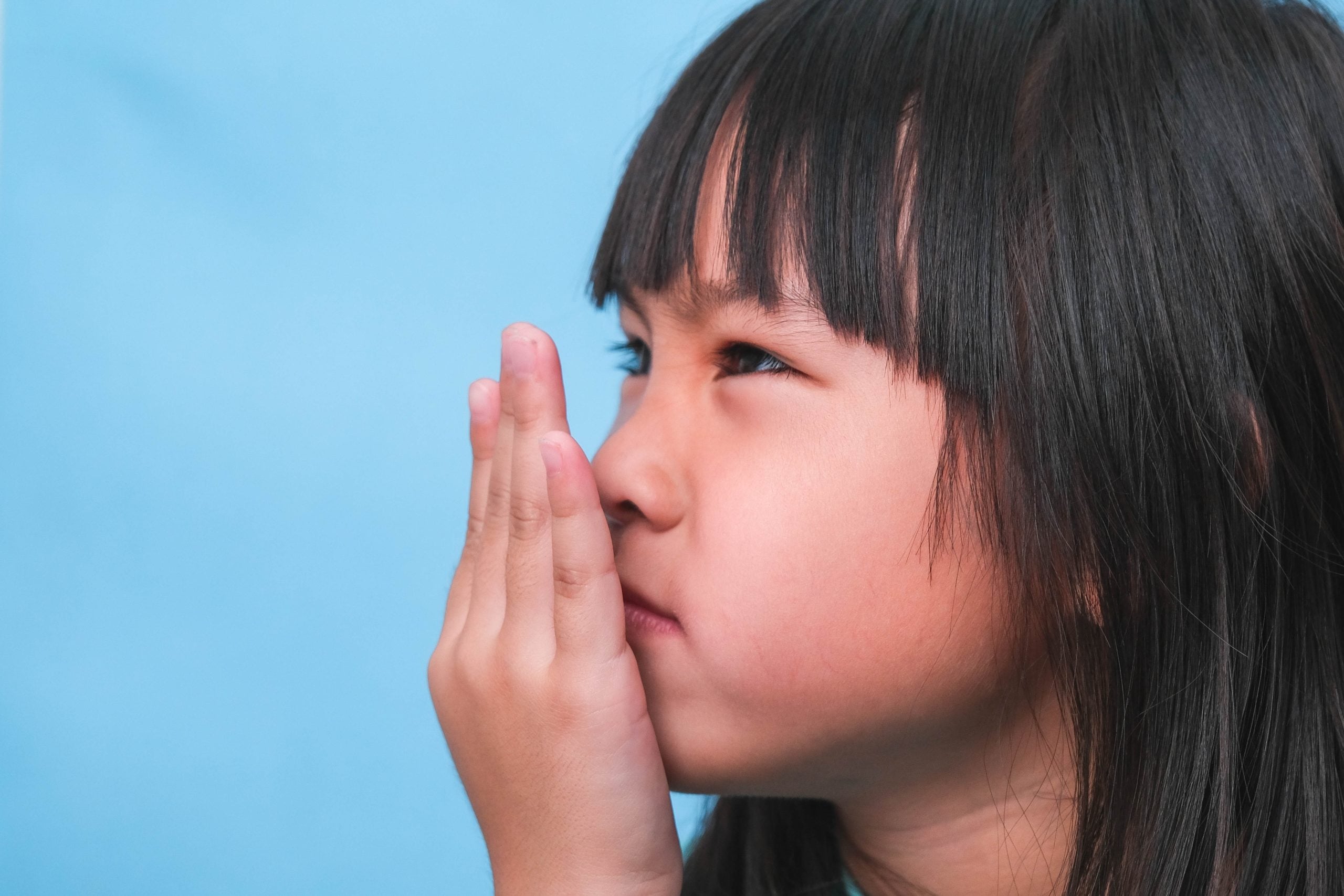 Little Asian girl covering her mouth to smell the bad breath. Child girl checking breath with her hands. Oral health problems or dental care concept.